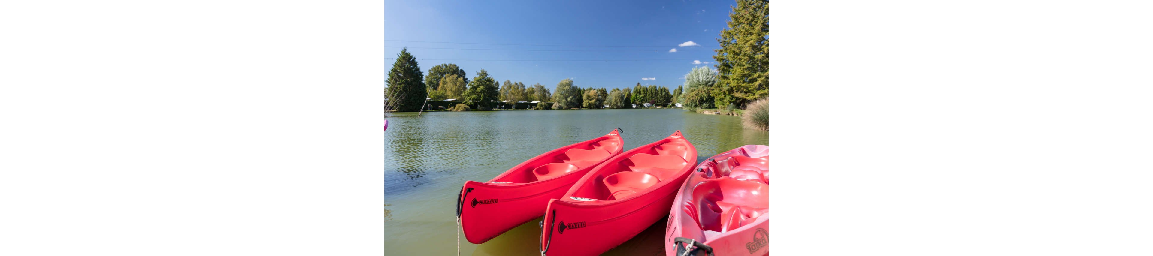 Three bright red kayaks rest at the edge of a calm lake under a clear blue sky. The water is smooth, reflecting the surrounding green trees and distant shoreline. In the background, a line of trees and small structures suggests a quiet lakeside campground or park. The scene feels peaceful and inviting, ideal for outdoor recreation and summer activities.