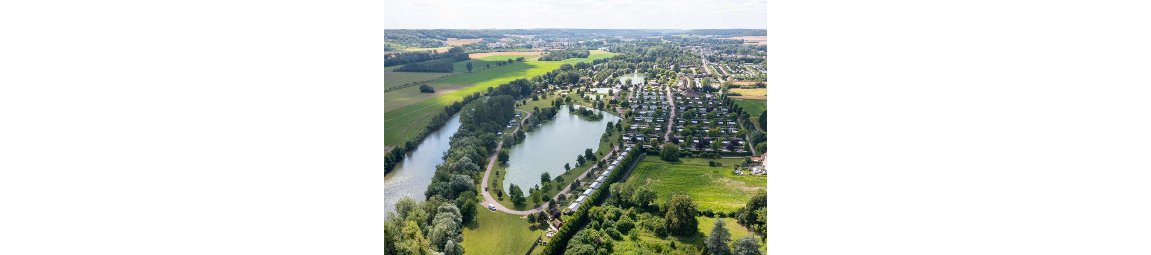 A large holiday park with rows of caravans and mobile homes is seen from above beside a winding river. Several lakes and ponds are surrounded by trees and open green fields. The setting appears rural, with farmland and a distant town visible on the horizon. The overall scene feels peaceful and spacious, emphasizing nature, water, and outdoor recreation.