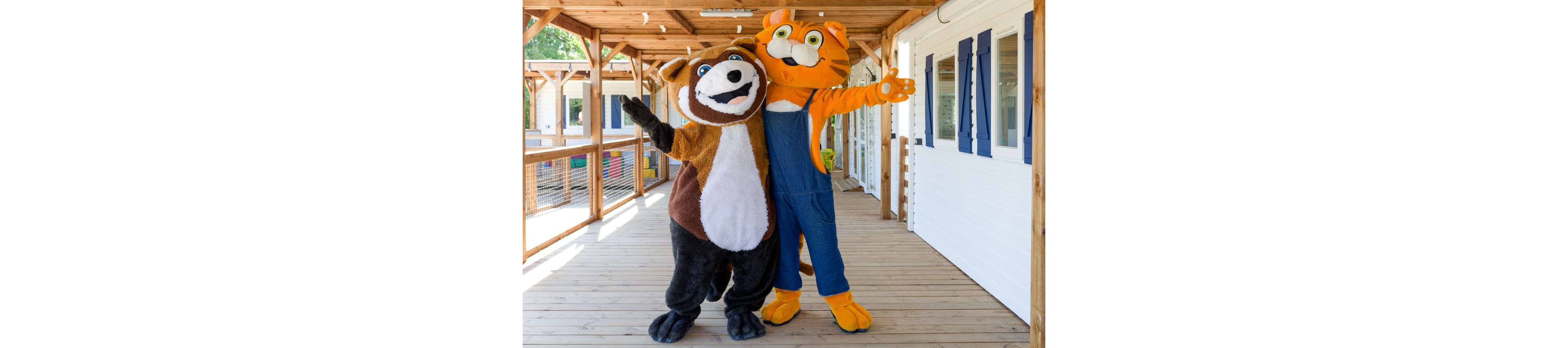 Two large animal mascots stand side by side on a wooden deck, posing with their arms around each other. One costume resembles a brown and white creature, while the other is an orange cat wearing blue overalls. They appear to be in a sunny outdoor setting with wooden railings and a light-colored building with blue shutters in the background. The bright daylight and playful characters create a fun, family-friendly atmosphere.