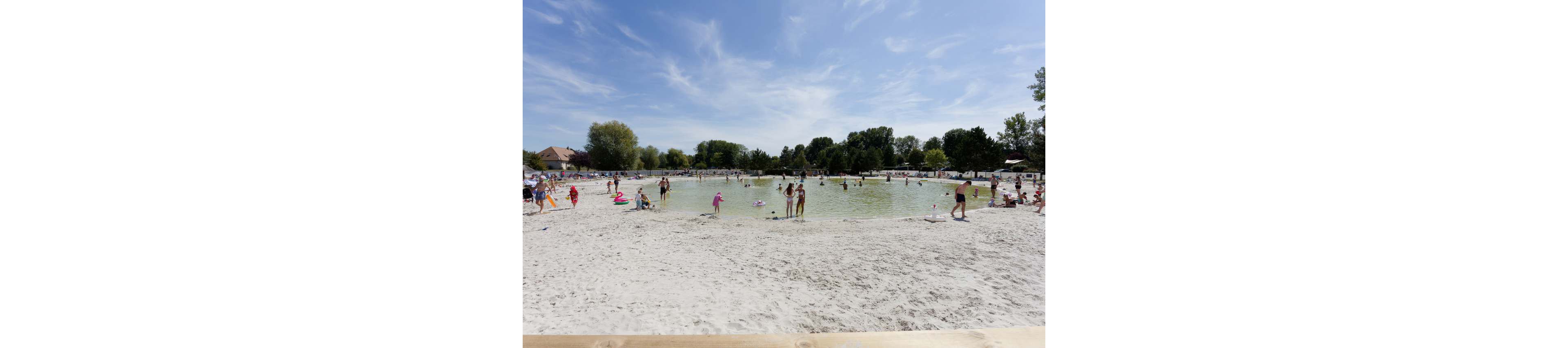 A sandy beach surrounds a shallow lakeside swimming area filled with people enjoying the water. Children and adults wade, swim, and play with inflatables under a bright blue sky with light clouds. Trees and small buildings line the background, suggesting a recreational park setting. The overall scene feels relaxed, summery, and family-friendly.