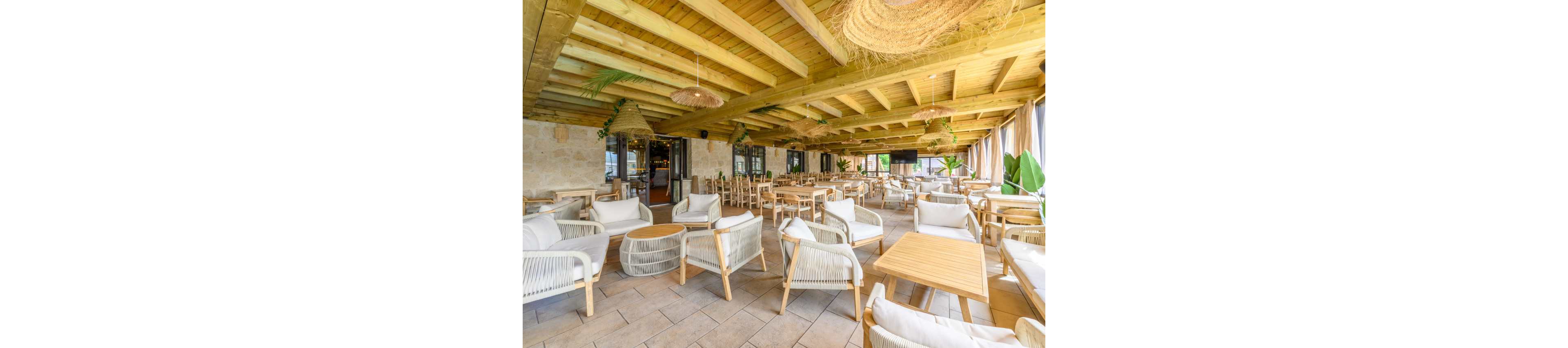 Spacious open-air restaurant terrace featuring light wooden tables and cushioned chairs. The setting includes a wooden beamed ceiling with woven pendant lights and natural stone walls. Large windows with curtains allow abundant daylight and views of greenery. The overall atmosphere feels relaxed, airy, and inviting for casual dining or social gatherings.