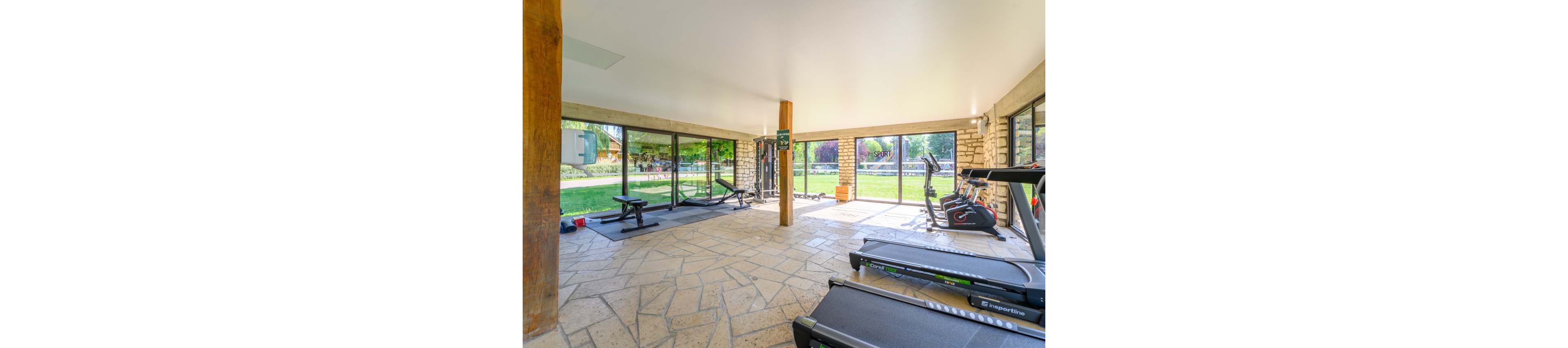 Spacious indoor gym room featuring treadmills, stationary bikes, and weight machines. Large floor-to-ceiling windows open onto a lush green garden, filling the space with natural light. The stone tile floor and wooden columns create a warm, contemporary look. No people are present, emphasizing the clean, organized workout environment.
