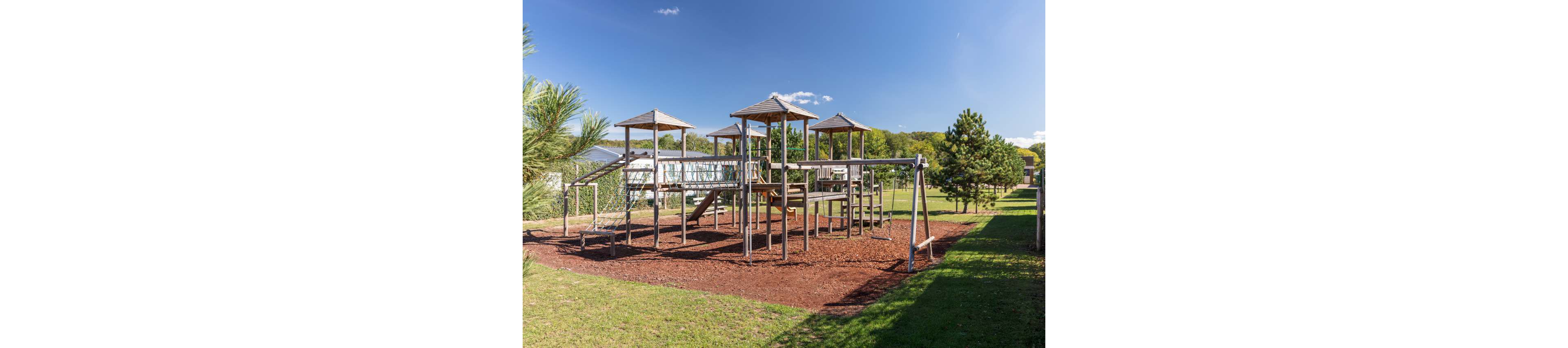 A large wooden playground structure stands on a bed of mulch in an open grassy park. Multiple towers, bridges, ladders, and swings create a varied play area for children. The scene is brightly lit under a clear blue sky with only a few small clouds. Trees and open fields surround the playground, giving it a peaceful, natural setting.