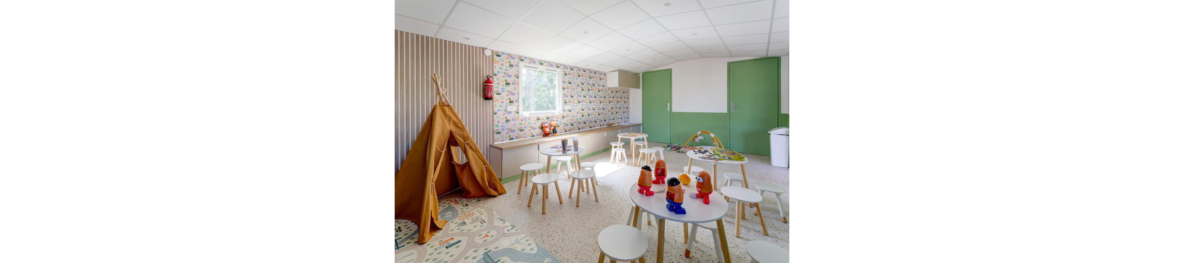 A colorful children's playroom features small round tables and stools arranged across a bright, spacious floor. Wooden toys and plastic figures are placed on the tables, while a play tent stands in one corner. The room has patterned wallpaper, green doors, and a large window letting in natural light. A wall-mounted fire extinguisher and a white trash bin are also visible, emphasizing a safe and tidy environment.