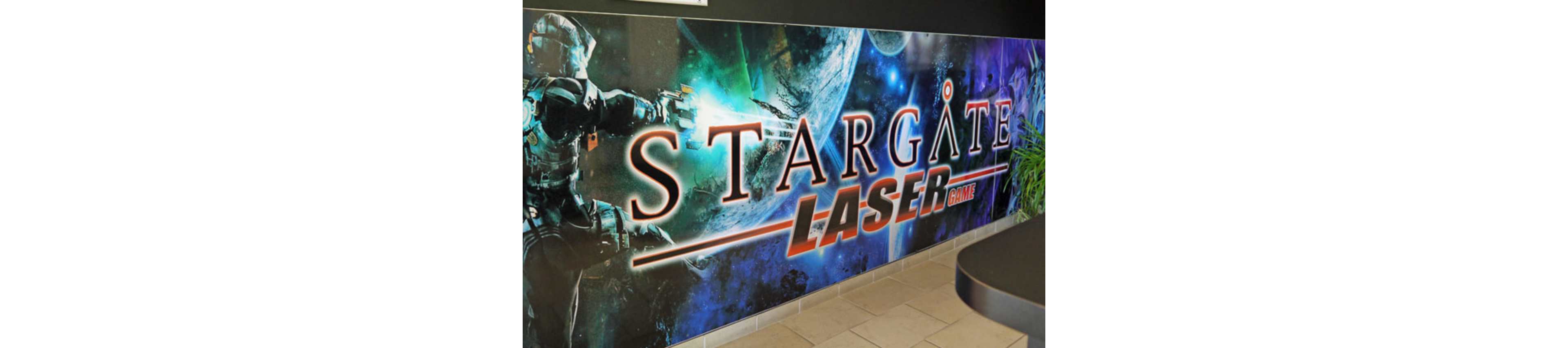 A colorful wall mural advertises a Stargate laser game experience. The artwork features a futuristic armored figure aiming a weapon in a deep space environment with planets and stars. Large bold text reads "STARGATE LASER GAME" across the center of the mural. The scene is located indoors, with tiled flooring and part of a counter visible in the foreground.