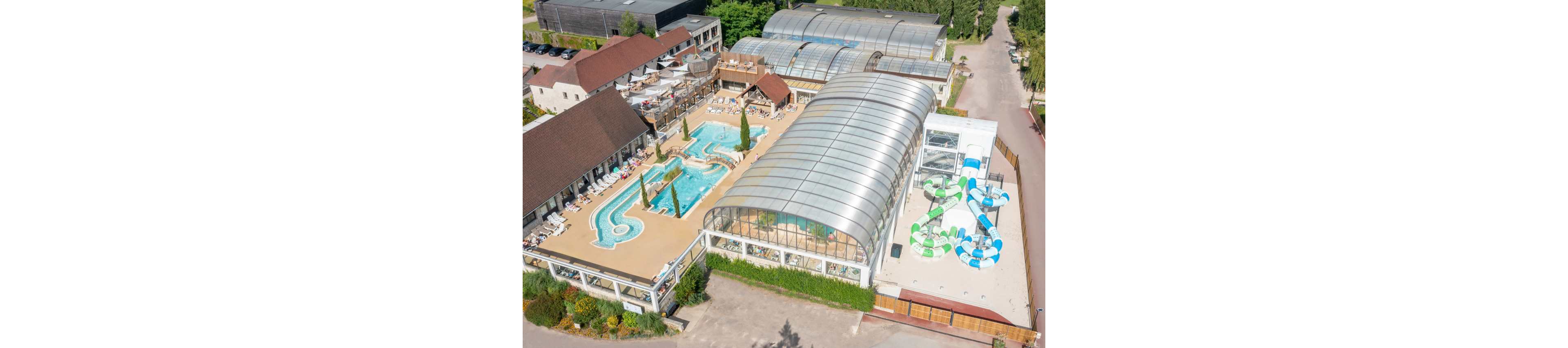 A large resort complex with multiple swimming pools and water slides is seen from an aerial perspective. The facility includes an outdoor pool area with sun loungers and a winding pool, alongside a large glass-covered indoor pool structure. Colorful spiral water slides extend from a tower into a separate pool zone. Surrounding buildings, greenery, and pathways frame the aquatic center, suggesting a family-friendly vacation destination.
