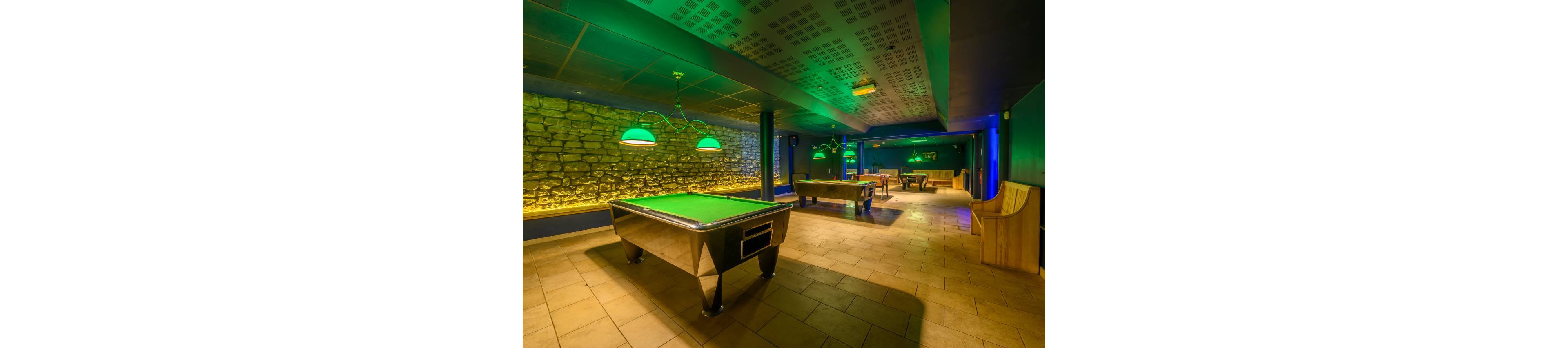 A spacious billiards room features a central pool table under bright green hanging lamps. The space has stone accent walls and warm yellow floor tiles, creating a cozy yet modern atmosphere. Additional pool and game tables are visible in the background, along with wooden seating areas. Green and yellow lighting adds a vibrant, nightlife feel to the recreational interior.