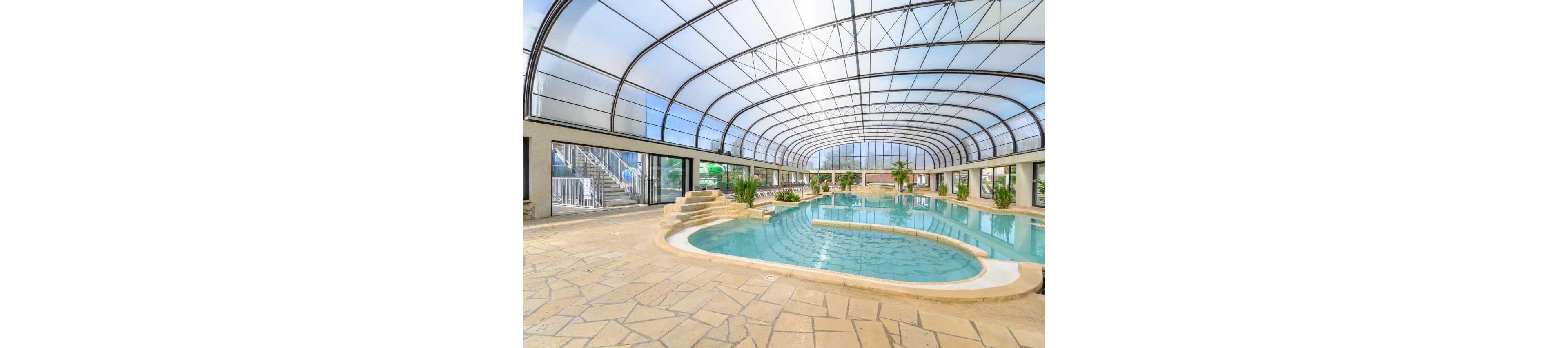 Large indoor swimming pool with a freeform shape sits beneath a high, curved glass roof. The bright space features stone-tiled flooring and built-in steps leading into the water. Green potted plants and poolside loungers add a resort-like atmosphere. Natural light floods the enclosure through expansive windows and transparent ceiling panels.