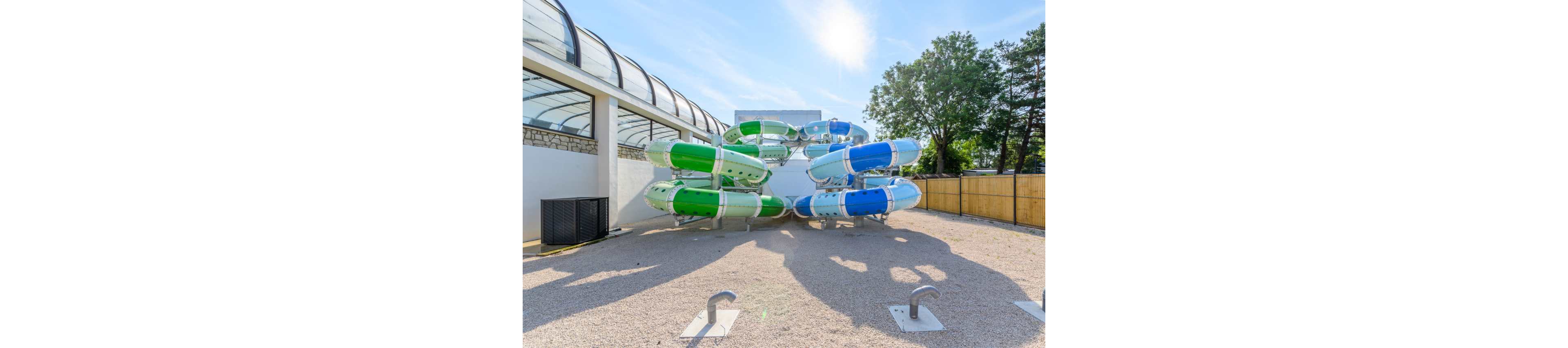 A cluster of enclosed water slides in blue, green, and white stands in an outdoor area of a water park. The slides emerge from a white building with a curved glass-covered walkway on one side. Metal fixtures and gravel cover the ground in the foreground, with no people present. Trees and a wooden fence line the background under a bright sunny sky.