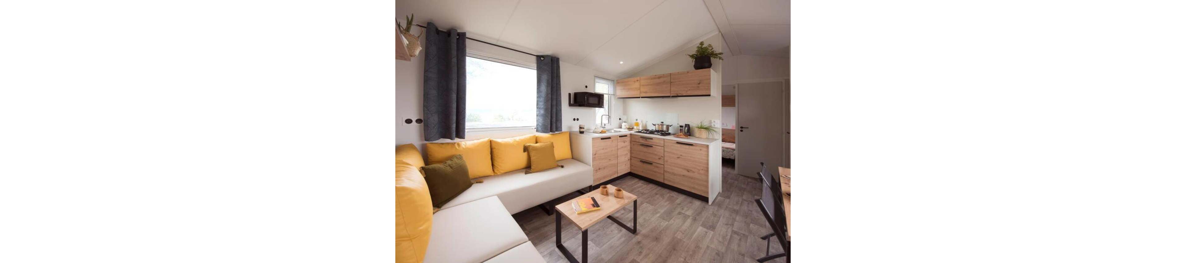 Compact open-plan living space features a modern kitchenette and cozy seating area. Light wood cabinets and neutral walls contrast with bright yellow cushions on an L-shaped sofa. A small coffee table and dining area complete the functional layout. Large window and ceiling lights provide ample natural and artificial light, while plants add a touch of greenery. A doorway in the background leads to a separate bedroom.