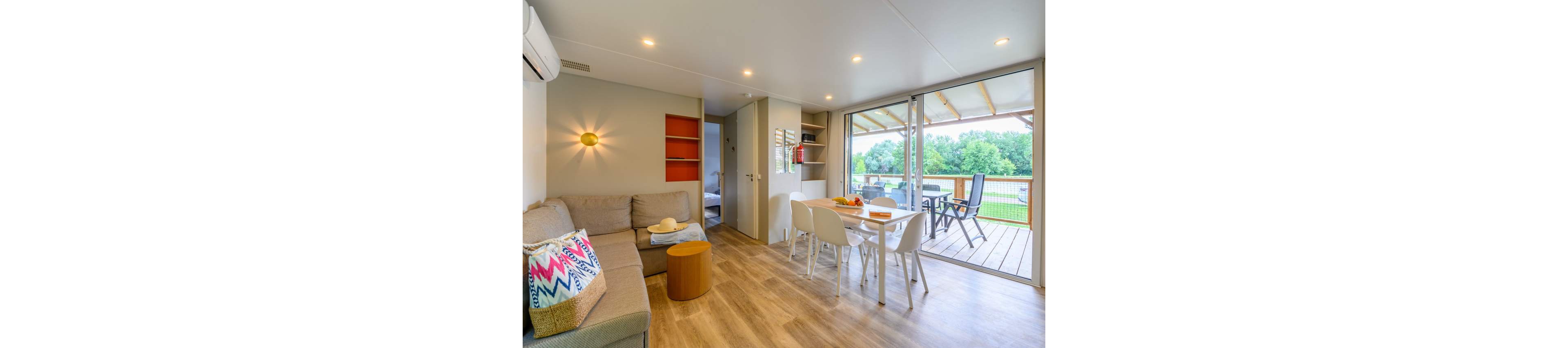 A bright modern cabin interior features a cozy sofa and compact dining table with white chairs. Large sliding glass doors open onto a covered wooden deck with outdoor seating and views of green trees. Warm wood flooring, recessed ceiling lights, and simple decor create a relaxed holiday atmosphere. The space appears tidy and inviting, ideal for a vacation rental or small family getaway.