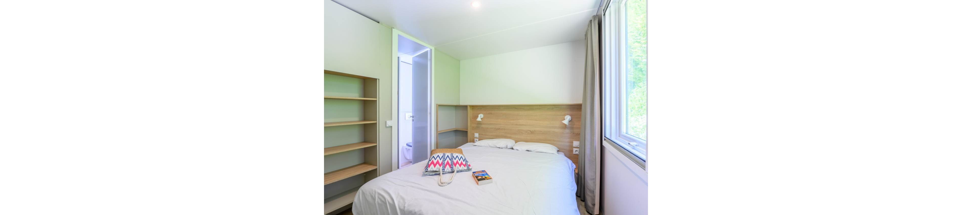A compact, modern bedroom features a neatly made double bed with a light wood headboard. Natural light streams in through a large window with beige curtains, illuminating the white walls and simple decor. An open shelving unit stands empty beside a doorway leading to a small bathroom. A colorful patterned tote bag and a paperback book rest on the bed, adding a casual, lived-in touch.