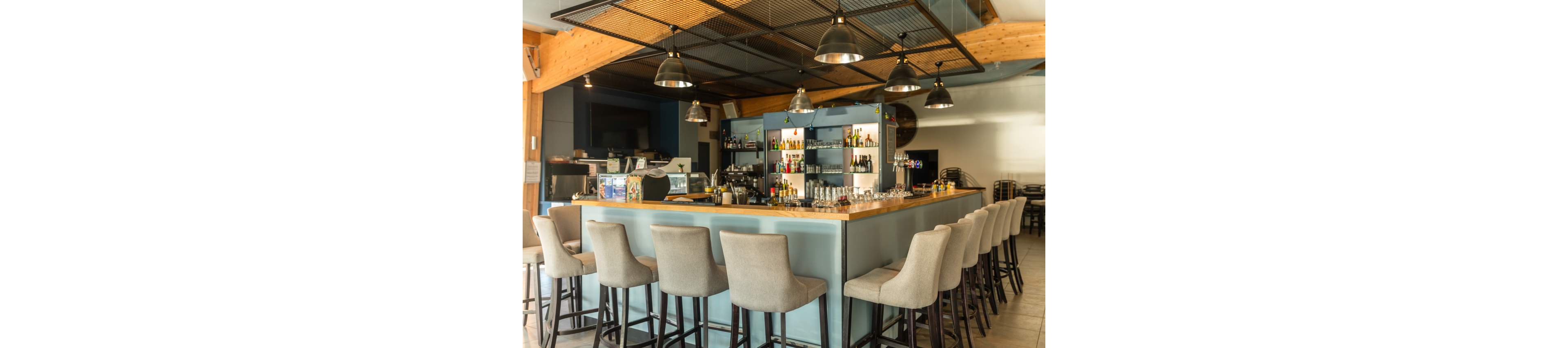 A contemporary bar interior features a square wooden counter surrounded by high upholstered stools. The bar shelves in the background display various bottles and glassware, with a coffee machine and other equipment on the counter. Industrial-style pendant lights hang from the ceiling, illuminating the clean and organized space. The setting appears ready for customers, emphasizing a relaxed yet stylish atmosphere.