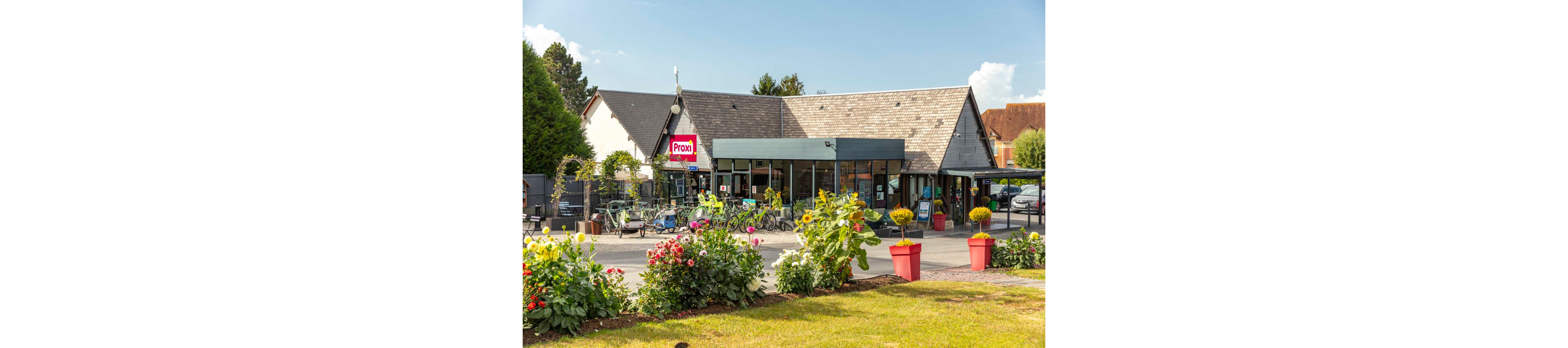A small Proxi supermarket stands behind a colorful flower bed on a sunny day. The building has a sloped roof and large glass windows at the entrance. Several bicycles are parked outside, suggesting customers visiting the store. Bright red planters and blooming flowers create a welcoming atmosphere in this quiet neighborhood setting.