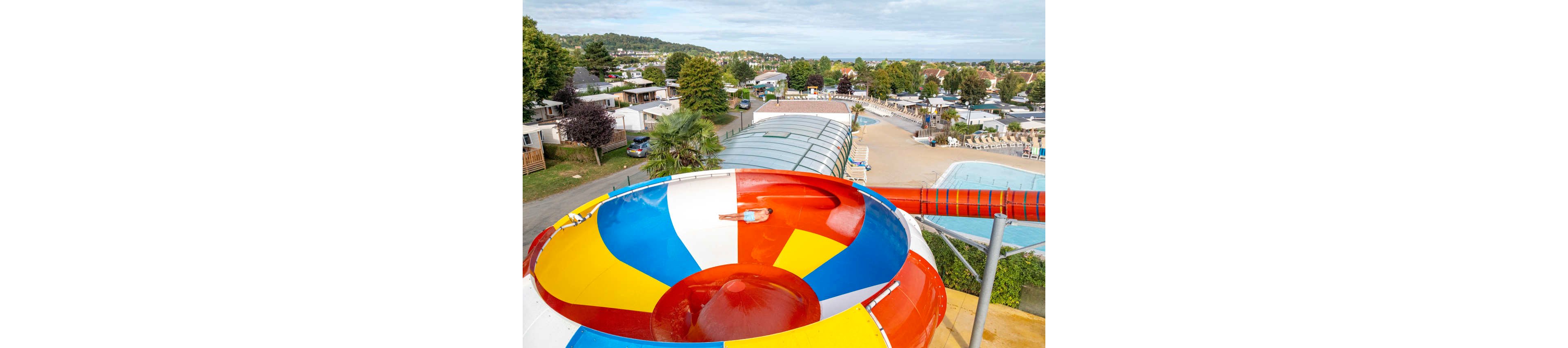 A large, colorful circular water slide dominates the foreground of an outdoor leisure complex. The slide features bright red, yellow, blue, and white sections and curves down toward a pool area. In the background, a swimming pool, sun loungers, and a covered pool structure are surrounded by trees and holiday cabins. The scene suggests a family-friendly vacation park on a clear day.