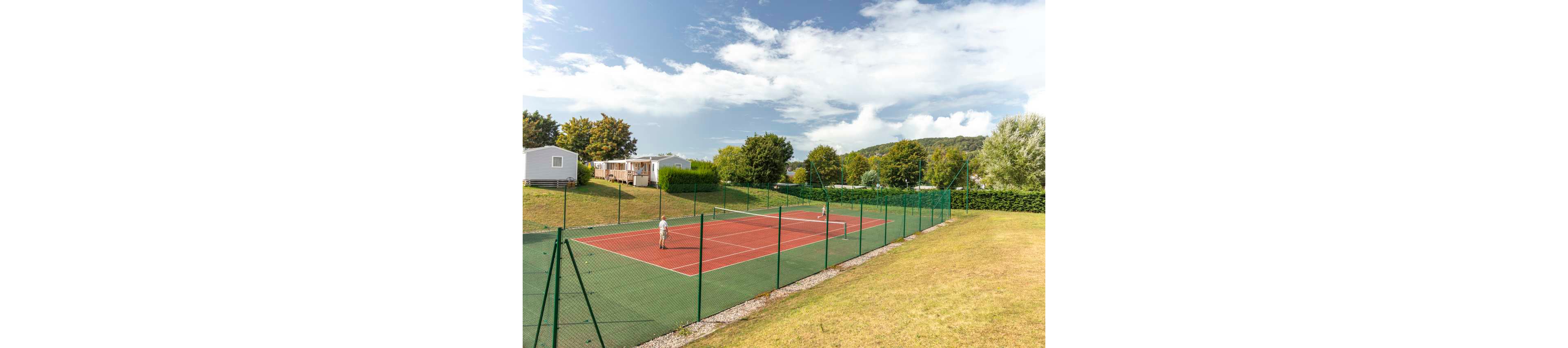 Two children are playing tennis on an outdoor court surrounded by green fencing. The court is located in a grassy area near a campsite with several mobile homes and trees in the background. The sky is partly cloudy, creating a bright and pleasant atmosphere. The scene suggests a relaxed holiday setting with recreational sports facilities.
