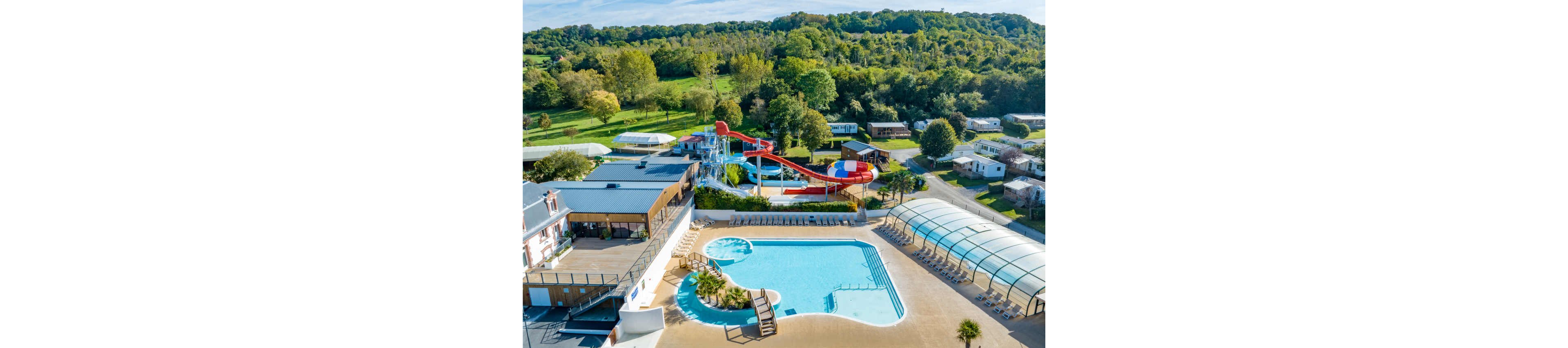A large outdoor swimming pool complex is shown from above within a countryside campsite. The scene features a main pool, a smaller circular pool, and a covered pool area with a transparent arched roof. Colorful water slides and play structures stand at the center, surrounded by sun loungers and low buildings. Mobile homes and caravans are scattered around the site, with lush green trees and rolling hills in the background under clear skies.
