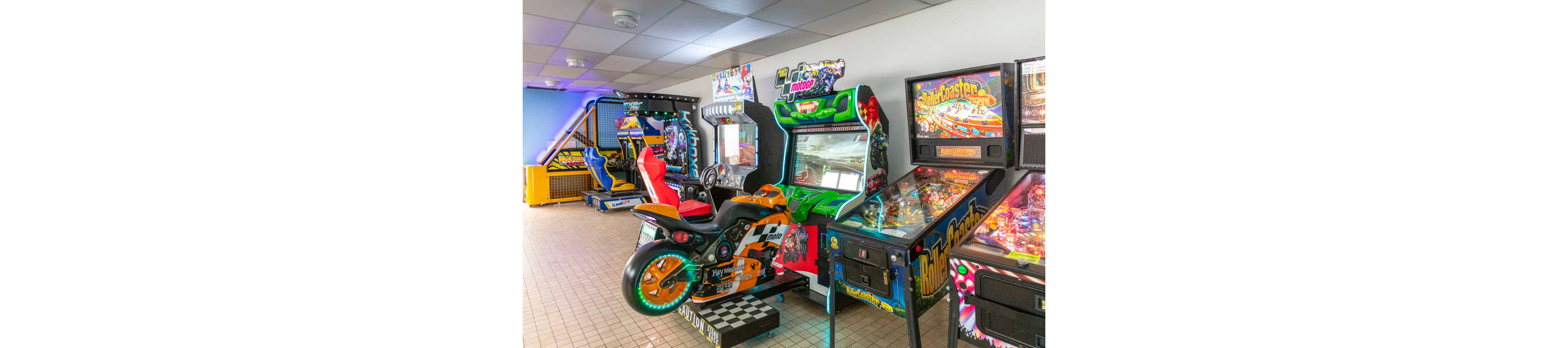A bright arcade room features a variety of gaming machines lined up against the wall. A motorcycle racing simulator stands prominently in the foreground, surrounded by pinball machines and other arcade cabinets. The tiled floor and overhead fluorescent lighting give the space a clean, casual atmosphere. Vivid colors, flashing lights, and illustrated game artwork create an energetic, playful environment.