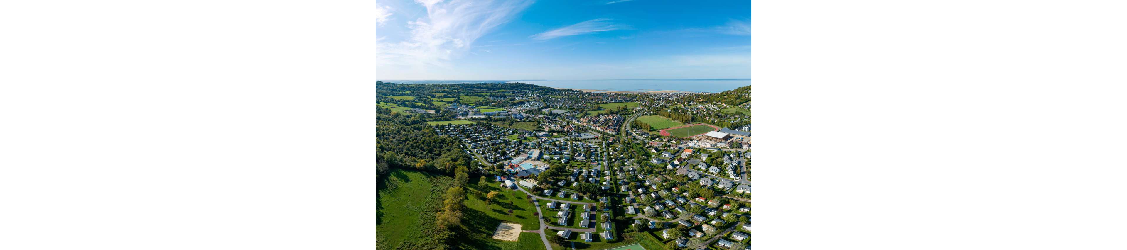 High-angle aerial photography captures a sprawling residential town near the coast. Numerous houses and mobile homes are arranged in neat rows, interspersed with trees and green spaces. A large sports field and several roads curve through the community, connecting different neighborhoods. In the distance, the shoreline and sea are visible under a bright blue sky with wispy clouds.