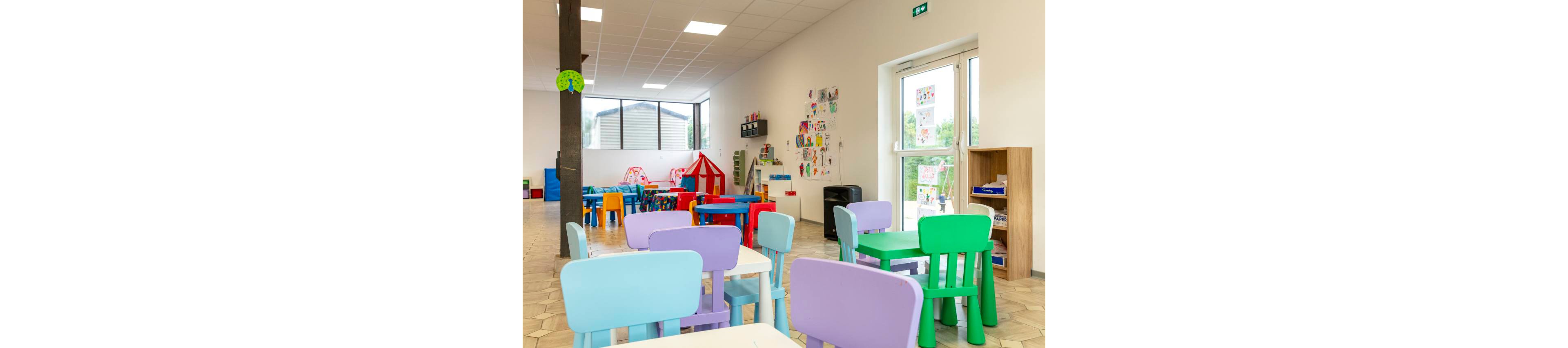 A spacious kindergarten classroom is filled with colorful plastic tables and chairs in the foreground. In the background, a play area includes small tables, toys, and a red-and-white play tent. Children's drawings and crafts are displayed on the walls near a large glass door that lets in natural light. The room feels cheerful and inviting, designed for young children to learn and play safely.