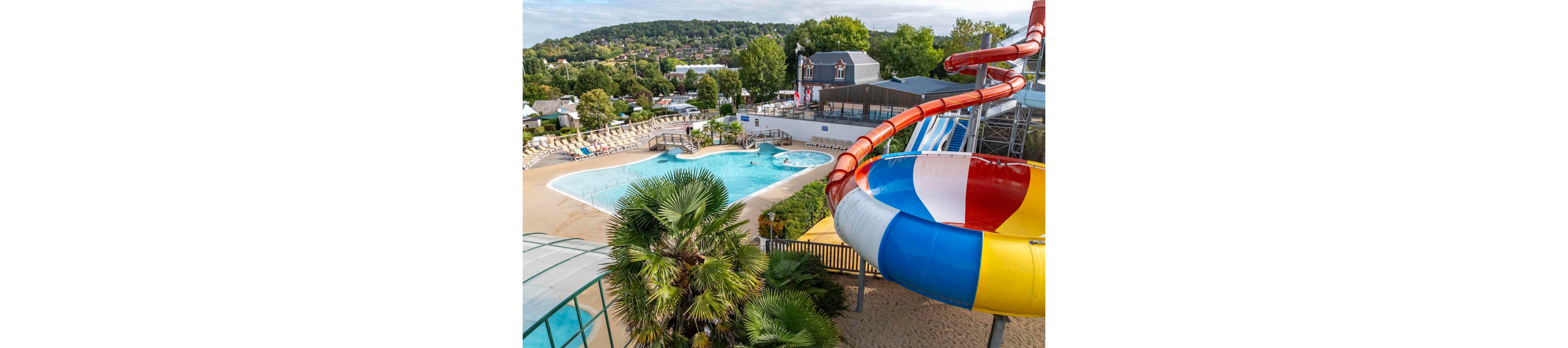 A large, colorful waterslide curves above an outdoor swimming pool at a resort. The pool area is surrounded by sun loungers, palm trees, and landscaped greenery. In the background, residential buildings and forested hills create a scenic setting. The scene suggests a family-friendly water park or campground on a bright day.