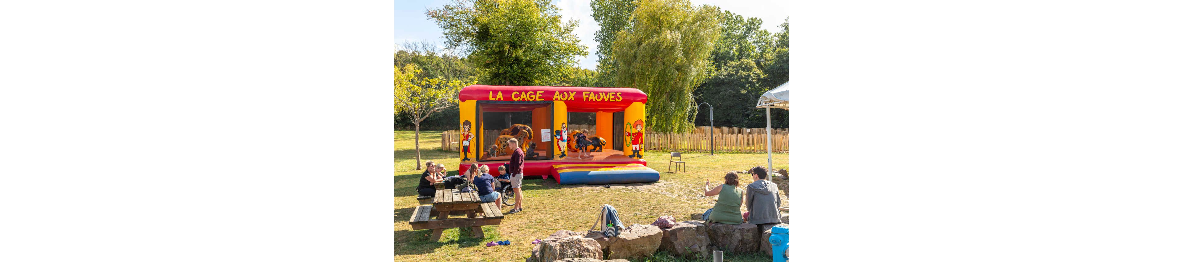 A bright inflatable bounce house labeled "LA CAGE AUX FAUVES" stands in a sunny park. Children jump and play inside the structure while adults sit nearby on benches and rocks. The scene is set on a grassy field with trees, a wooden fence, and a relaxed picnic atmosphere. Warm daylight and vivid primary colors create a cheerful, family-friendly mood.