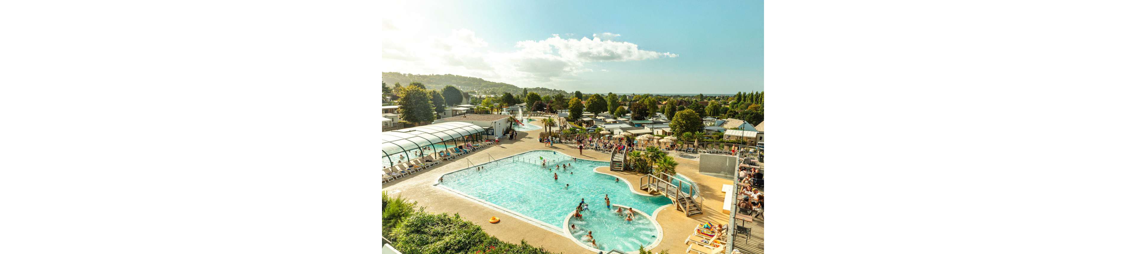 An elevated view shows a lively holiday resort with multiple swimming pools filled with people. Families and children play in the main outdoor pool while others relax on sun loungers around the deck. A partially covered pool structure and water slide area are visible in the background. The scene is set on a sunny day with green hills, trees, and numerous vacation cabins surrounding the aquatic area.