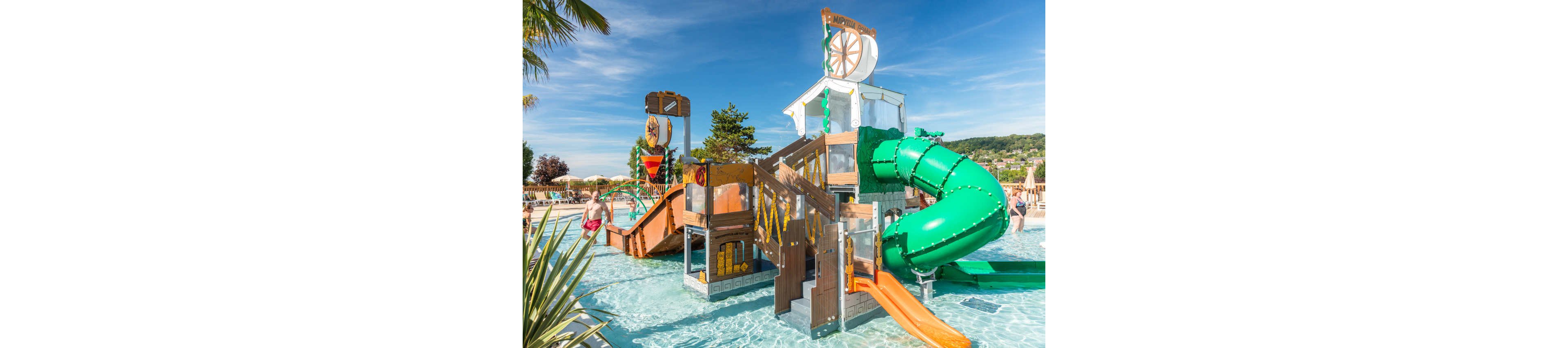 A vibrant water playground features multiple slides and climbing structures in a shallow pool. Children and adults enjoy the attractions under a clear blue sky on a sunny day. The play area includes a large green tube slide, smaller open slides, and themed decorative elements. Lush greenery and distant hills frame the family-friendly aquatic setting.