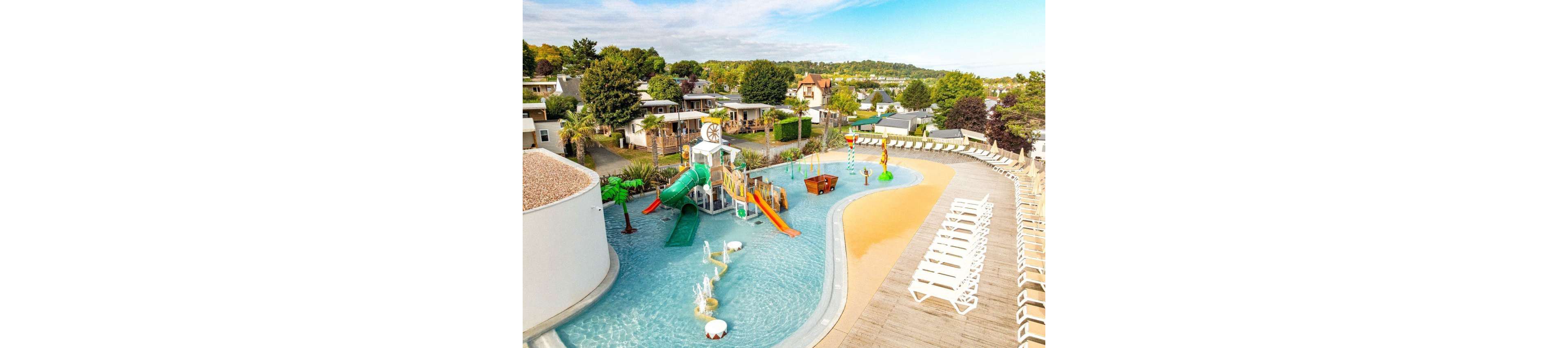 An outdoor resort-style swimming pool features a colorful children's water playground with slides and climbing structures. The pool curves around a deck lined with rows of white lounge chairs for sunbathing. Surrounding the pool area are holiday homes, trees, and landscaped greenery, creating a relaxed vacation atmosphere. The scene is bright and sunny, emphasizing leisure and family-friendly fun.