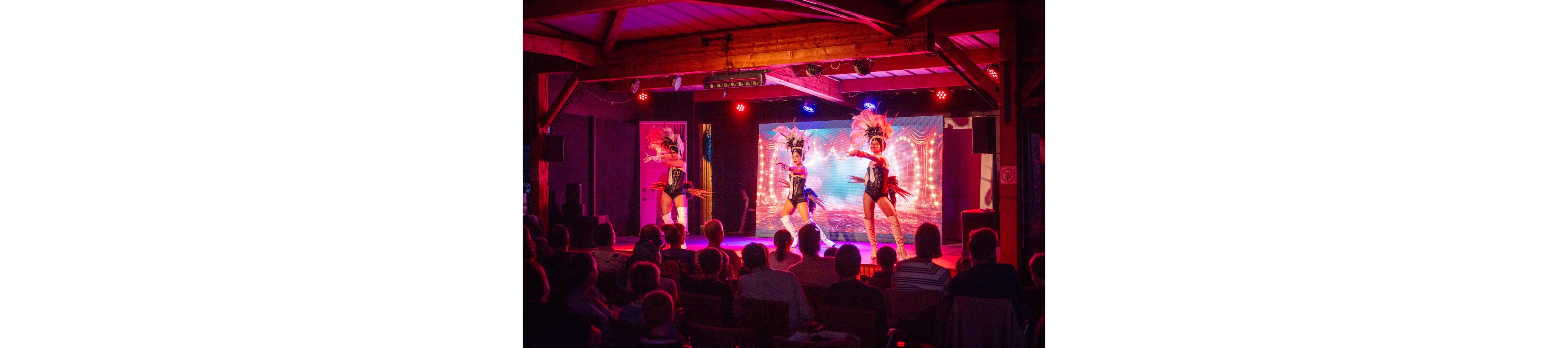 Three cabaret-style dancers perform on a brightly lit stage in front of a seated audience. The performers wear elaborate feathered headdresses and sparkling costumes, highlighted by vivid red and blue stage lighting. The background features a decorative curtain and illuminated backdrop that enhance the theatrical atmosphere. Audience silhouettes in the foreground emphasize the live entertainment setting and nighttime mood.