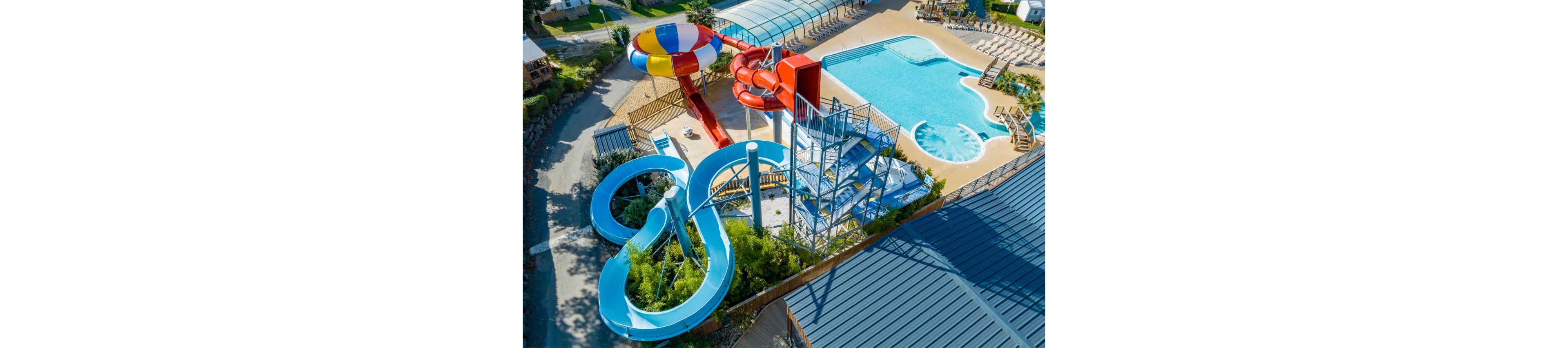 A bright aerial view showcases a water park with multiple twisting slides and a large swimming pool. The scene includes blue open slides, enclosed red tube slides, and a multicolored funnel slide structure. Sun loungers line the poolside area, suggesting a relaxing vacation setting. The overall atmosphere is sunny, fun, and family-friendly.