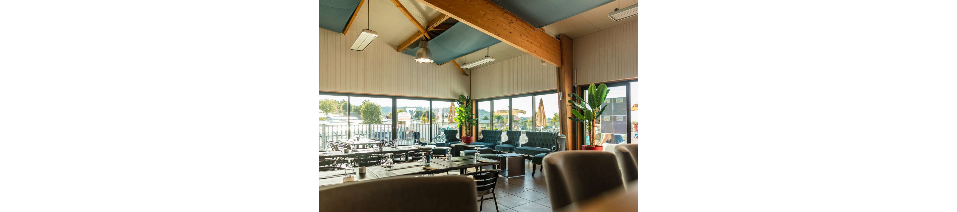 A spacious cafe interior features neatly arranged tables and chairs in front of floor-to-ceiling windows. Comfortable sofas and armchairs create a lounge area near the glass walls, with views of an outdoor terrace and surrounding landscape. Warm wooden beams, tiled flooring, and potted plants add a cozy yet contemporary atmosphere. The space is empty of people, emphasizing its clean, inviting setup for dining or social gatherings.