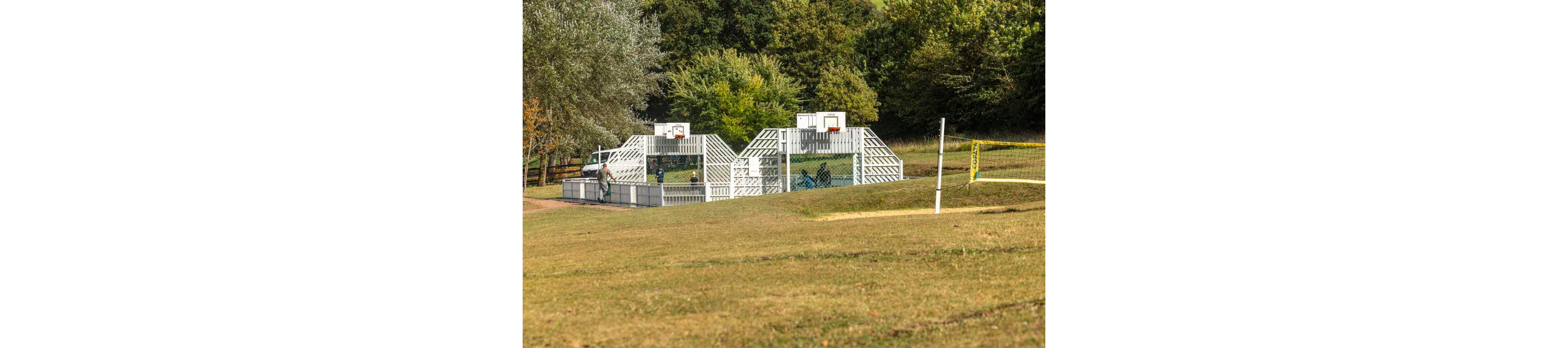 An outdoor multi-sport court with basketball hoops stands in the middle of a grassy park. A few people are gathered inside the fenced court area, suggesting casual play or practice. The surrounding landscape features dry grass in the foreground and dense green trees in the background. A volleyball or tennis net is set up nearby, emphasizing the recreational nature of the setting.