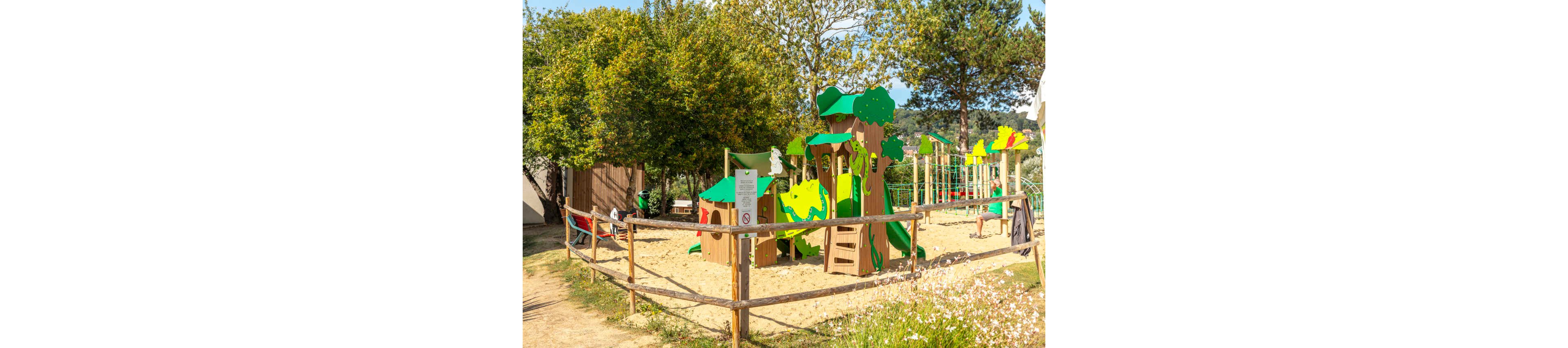 A bright outdoor playground with nature-themed equipment stands in a sandy play area. Wooden structures with green and yellow panels resemble trees and animals, surrounded by a simple wooden fence. Lush trees and greenery frame the scene under a clear, sunny sky. A sign is posted near the entrance, and a few people are visible in the background enjoying the park.