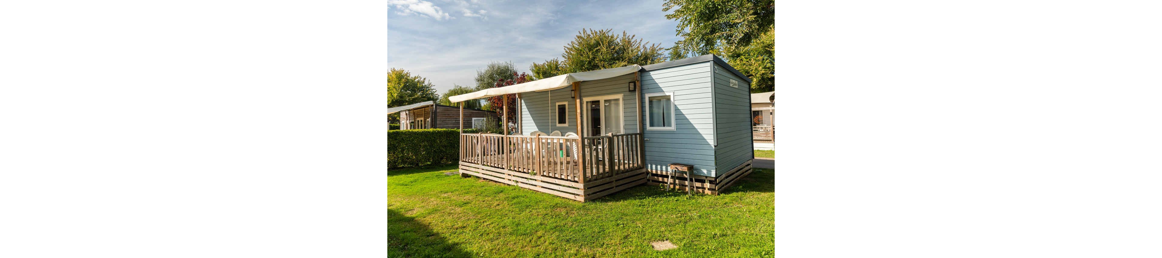 A light blue mobile home with a spacious wooden terrace sits on a well-kept grassy plot. White outdoor chairs and a table are arranged under a large awning that provides shade. The home is surrounded by hedges, trees, and neighboring cabins in the background. Bright daylight and clear skies create a relaxed holiday atmosphere in this small vacation residence.