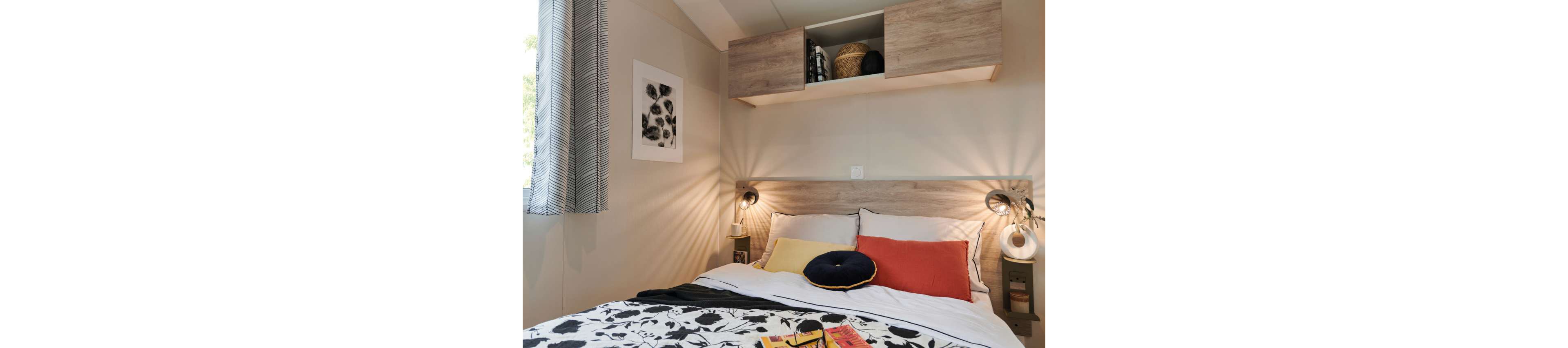 A neatly made bed with patterned black and white bedding is the focal point of this compact modern bedroom. Colorful accent pillows in yellow, navy, and red add warmth against a light wood headboard. Wall-mounted lamps provide soft lighting on either side of the bed, while open overhead cabinets display baskets and decor. A framed botanical print and striped curtains complete the cozy, contemporary look.