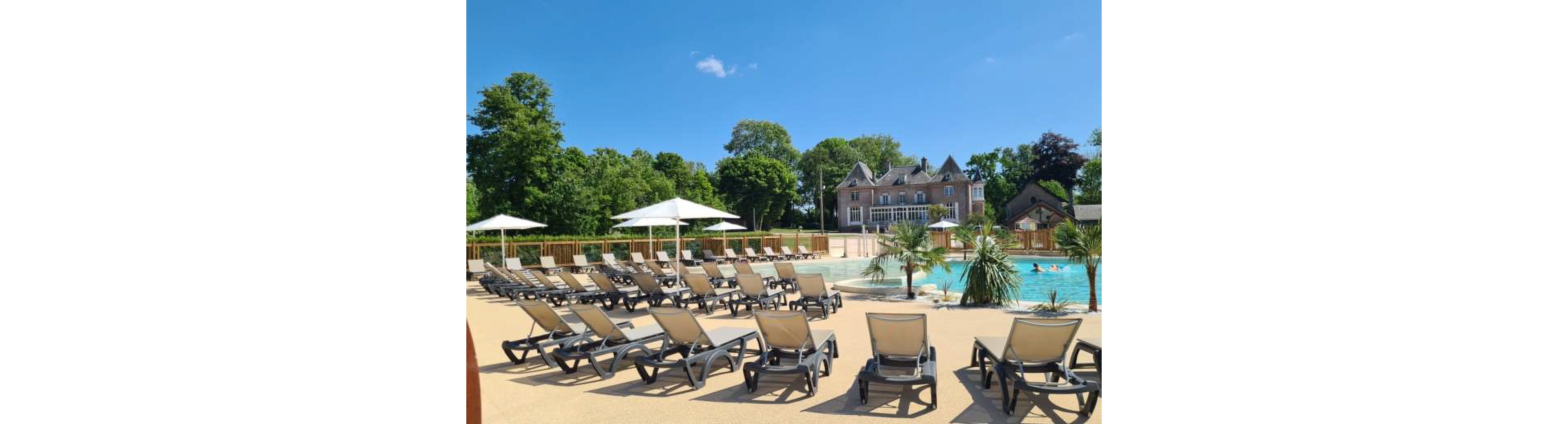 A large outdoor swimming pool is surrounded by rows of empty sun loungers under a clear blue sky. White parasols provide shade along the poolside terrace, while a few people swim and play in the water. In the background, a grand house or villa stands among tall green trees, giving the scene an upscale resort feel. Small palm trees and clean paved surfaces enhance the relaxed, vacation atmosphere.