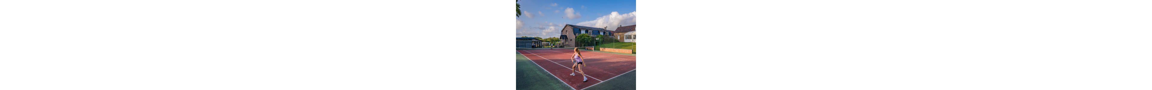 A young girl prepares to hit a tennis ball on an outdoor court. She stands near the baseline in an athletic stance, holding a racket with both hands. Behind her, residential-style buildings and greenery frame the scene under a bright blue sky with scattered clouds. The warm sunlight and open setting create a relaxed, active atmosphere.