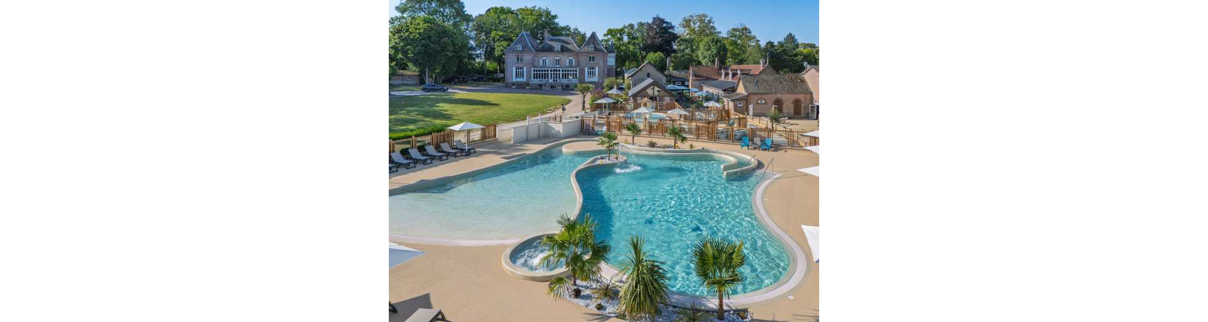 A large, curving outdoor swimming pool dominates the foreground of this resort scene. Sun loungers line the pool deck, with umbrellas and seating areas arranged around the water. In the background, a grand country house and smaller rustic buildings overlook the pool area. Lush green lawns, mature trees, and a few decorative palm plants create a relaxed holiday atmosphere under clear skies.