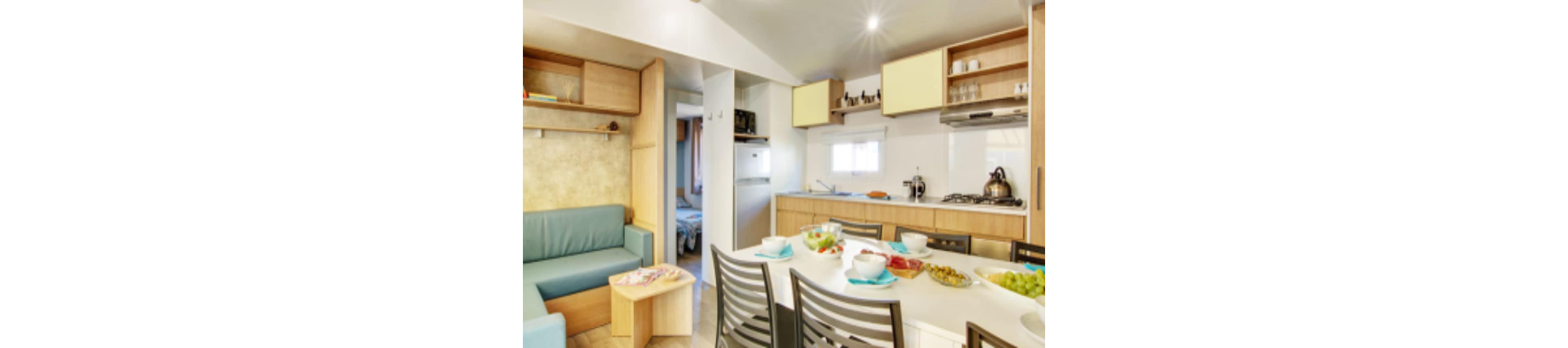 A compact modern kitchen opens into a cozy dining and living area. A white table is set with bowls, plates, and fresh fruit, surrounded by metal chairs. Light wood cabinetry, open shelves with glassware, and a gas stove with a kettle create a functional cooking space. A small sofa and side table sit nearby, with a doorway leading to a bedroom in the background.