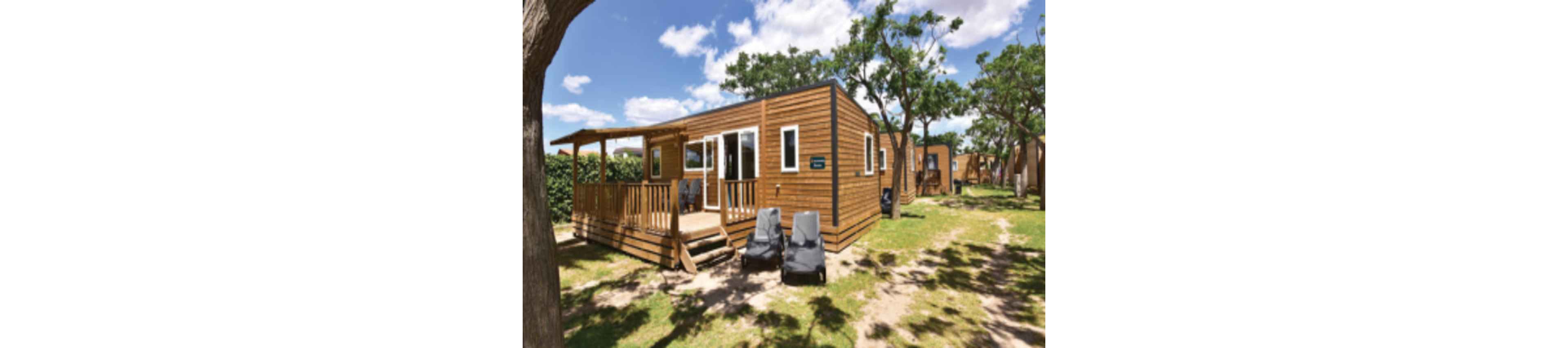 A modern wooden cabin stands in a sunny outdoor setting surrounded by trees and grass. The cabin features large windows, a small front porch with railing, and steps leading down to the yard. Two reclining lounge chairs are placed on the lawn, suggesting a relaxing vacation atmosphere. Other similar cabins are visible in the background, indicating a holiday park or campground environment.