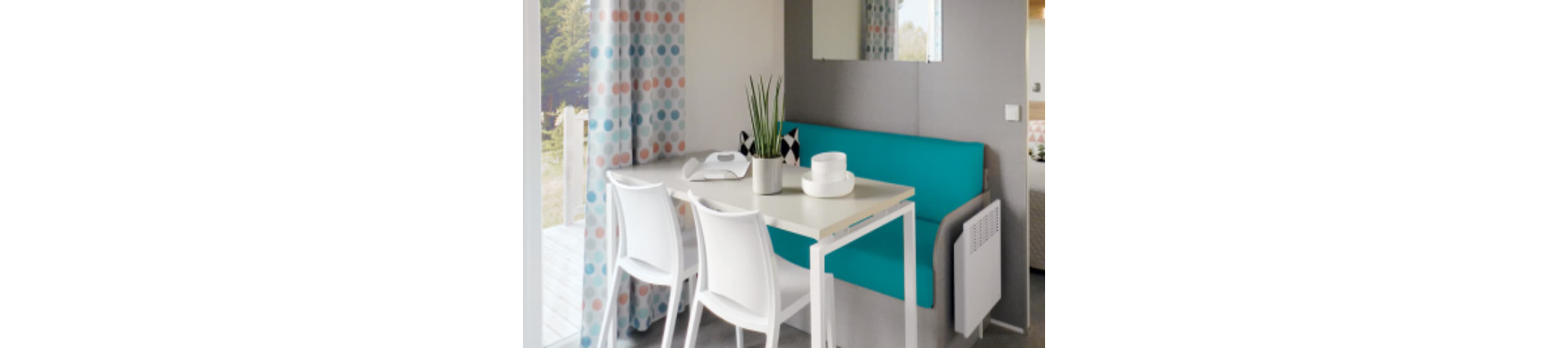 A compact dining nook features a white table with two white chairs and a teal upholstered bench. The space is decorated in a modern style with a potted plant and simple white tableware on the tabletop. Colorful polka dot curtains frame a doorway leading to an outdoor area, while a mirror and neutral walls keep the room feeling open. Soft natural light enhances the clean, minimalist design of this small interior.