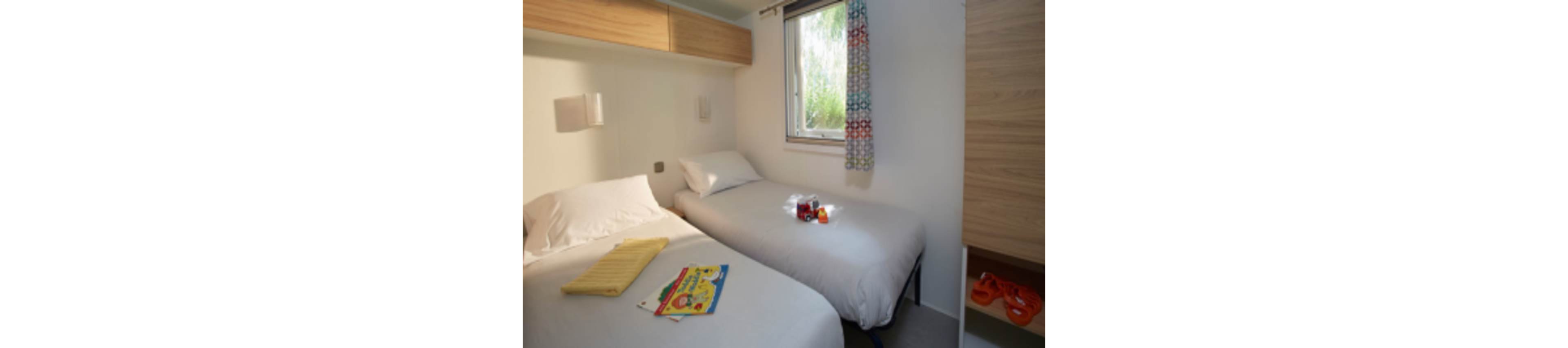 A small, bright bedroom features two neatly made single beds separated by a narrow gap. Daylight enters through a window with patterned curtains, illuminating the simple modern furnishings and overhead storage. A magazine and folded yellow towel rest on one bed, while a tray with drinks and snacks sits on the other. The overall atmosphere is clean, casual, and inviting, suggesting a vacation rental or compact guest room.