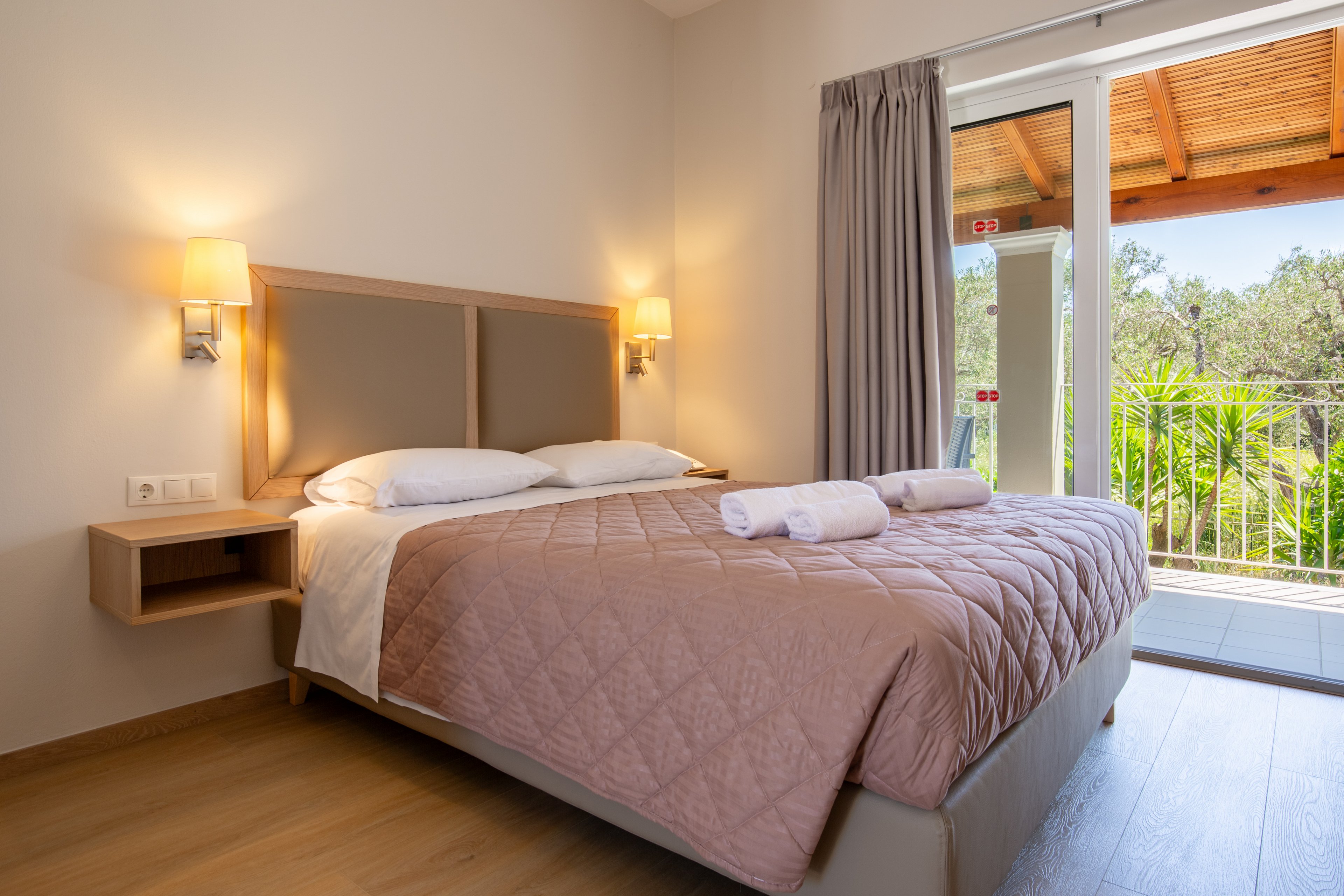 A neatly made double bed with a quilted beige coverlet stands in a modern hotel bedroom. Wall-mounted bedside lamps cast a warm light on the neutral walls and wooden floor. Fresh towels are folded on the bed, and a large glass door opens to a balcony with greenery outside. The room feels airy and inviting, with simple contemporary furnishings and soft natural light.