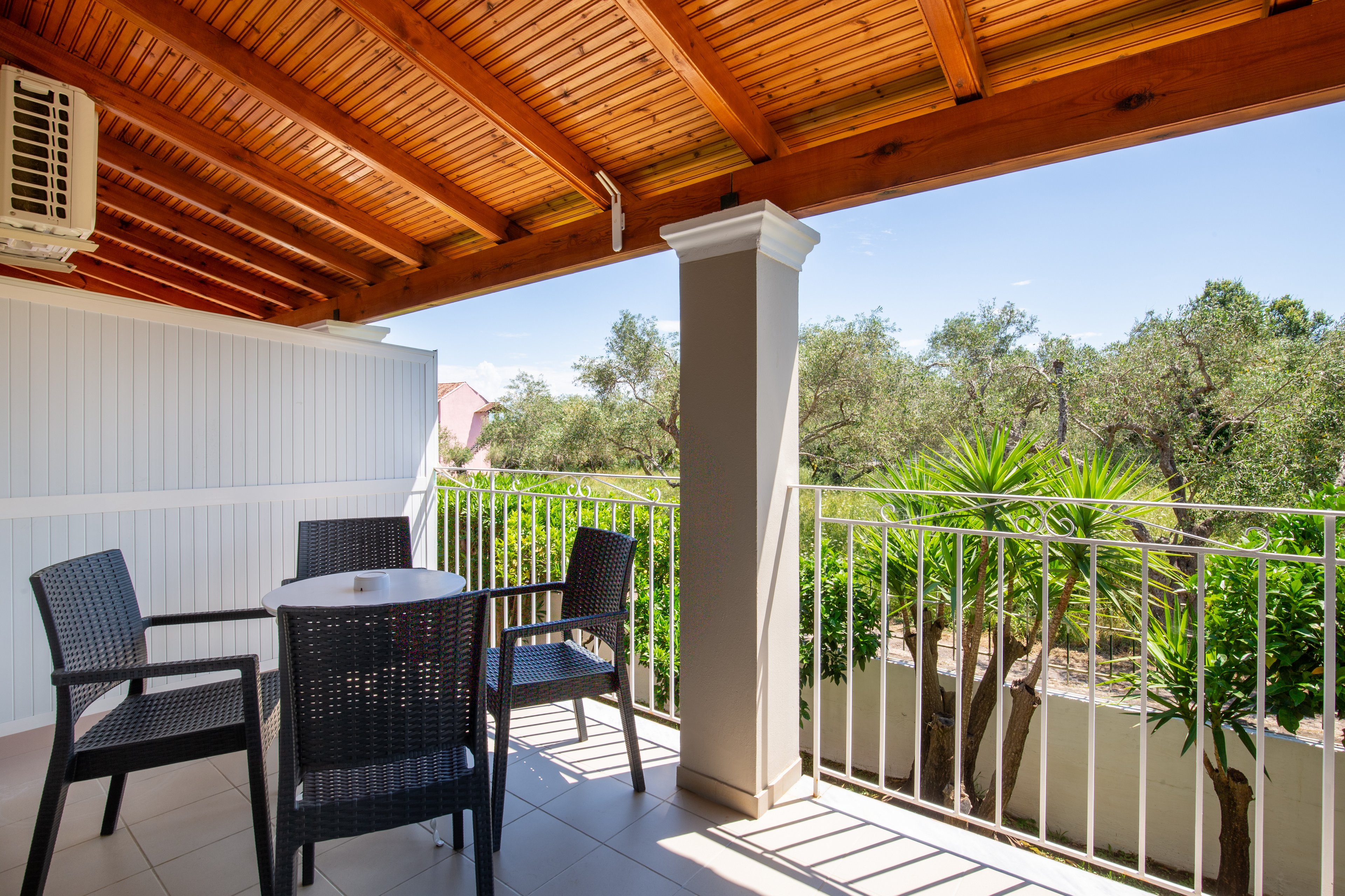 An outdoor balcony terrace features a small round table with four dark wicker chairs under a wooden pergola roof. The space is partially enclosed with white paneling and supported by a central column. A white metal railing frames the edge of the balcony, overlooking lush green trees and plants. Bright daylight and clear blue sky create a warm, inviting atmosphere.