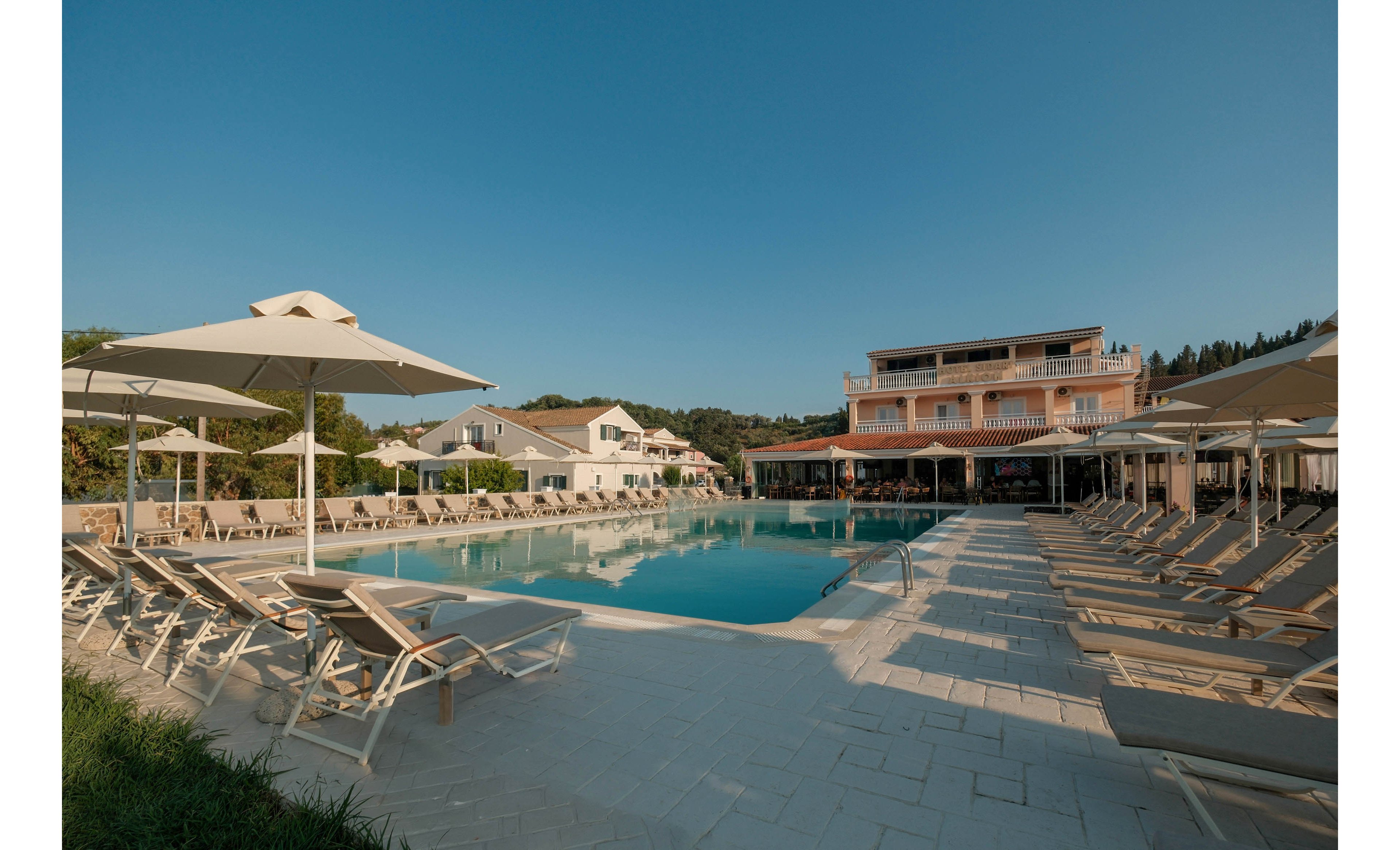 An outdoor resort swimming pool is surrounded by rows of sun loungers and large umbrellas under a clear blue sky. A multi-story hotel building and smaller villas stand in the background, framed by trees and greenery. The scene is calm and inviting, suggesting a relaxing vacation atmosphere in warm daylight. Clean stone paving and reflections in the water emphasize the neat, modern design of the pool area.