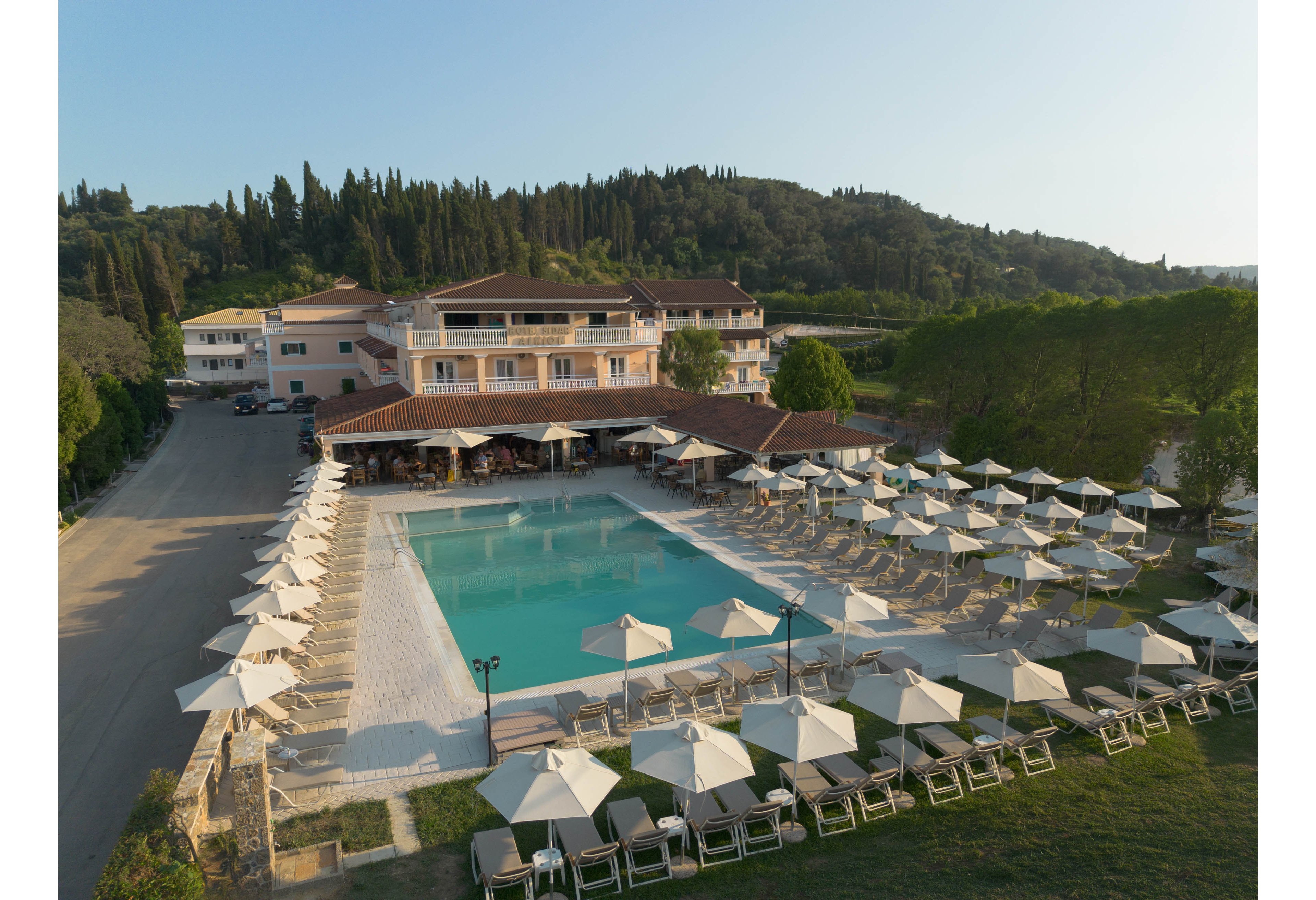 A large outdoor swimming pool is surrounded by rows of sun loungers and umbrellas at a resort complex. The scene is captured from above, showing the main building, poolside bar area, and adjacent road. Lush green trees and hills form a natural backdrop, creating a tranquil holiday atmosphere. The clear water and orderly arrangement of seating emphasize relaxation and leisure in a warm, sunny setting.