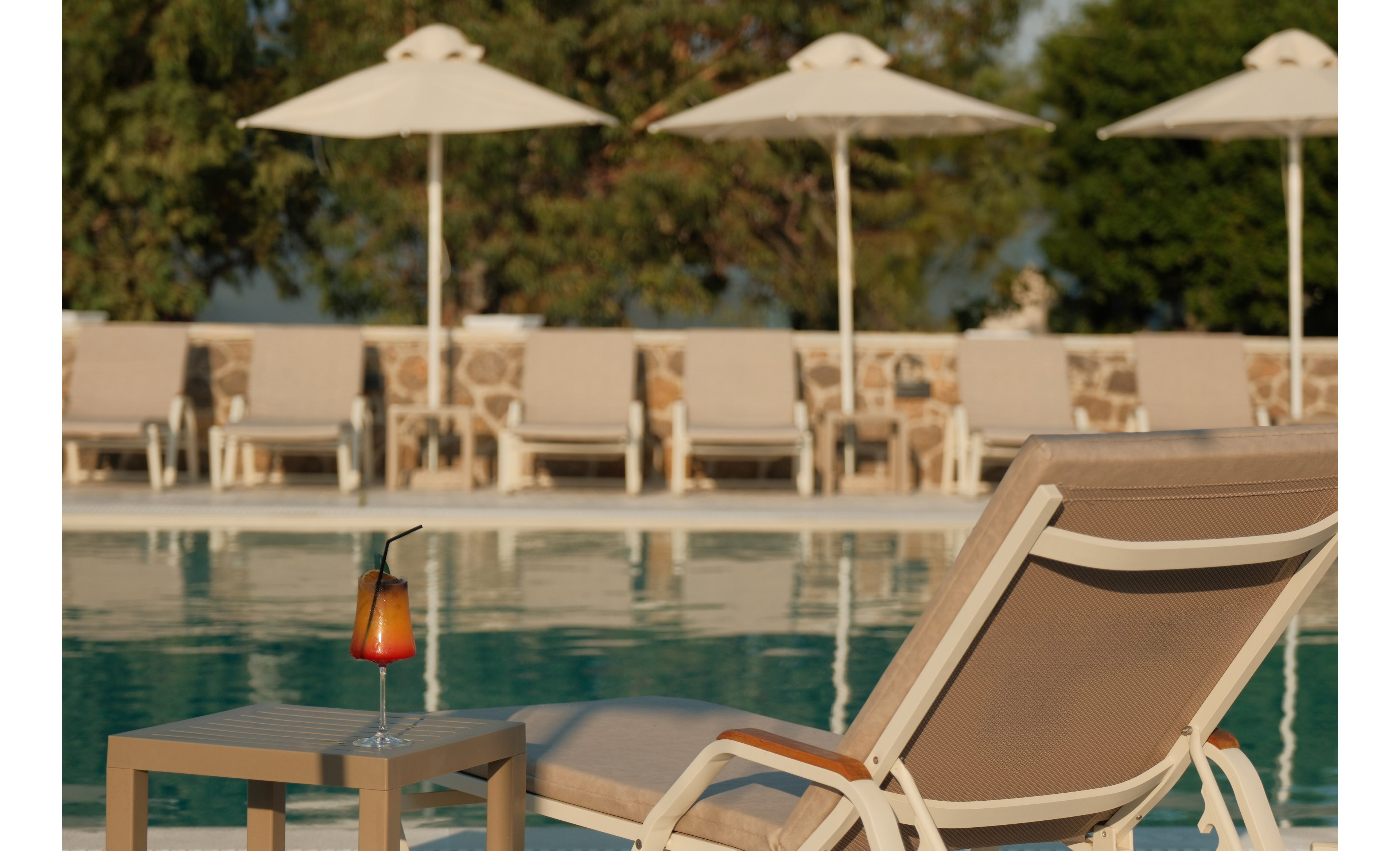 A cushioned lounge chair sits beside a clear outdoor swimming pool, with a colorful layered cocktail on a small side table. In the background, multiple sun loungers and large white umbrellas line the pool deck, suggesting a resort or hotel setting. The scene is bathed in warm sunlight, creating a relaxed vacation atmosphere with reflections on the calm water. Trees and a stone wall add natural and architectural elements to the tranquil environment.