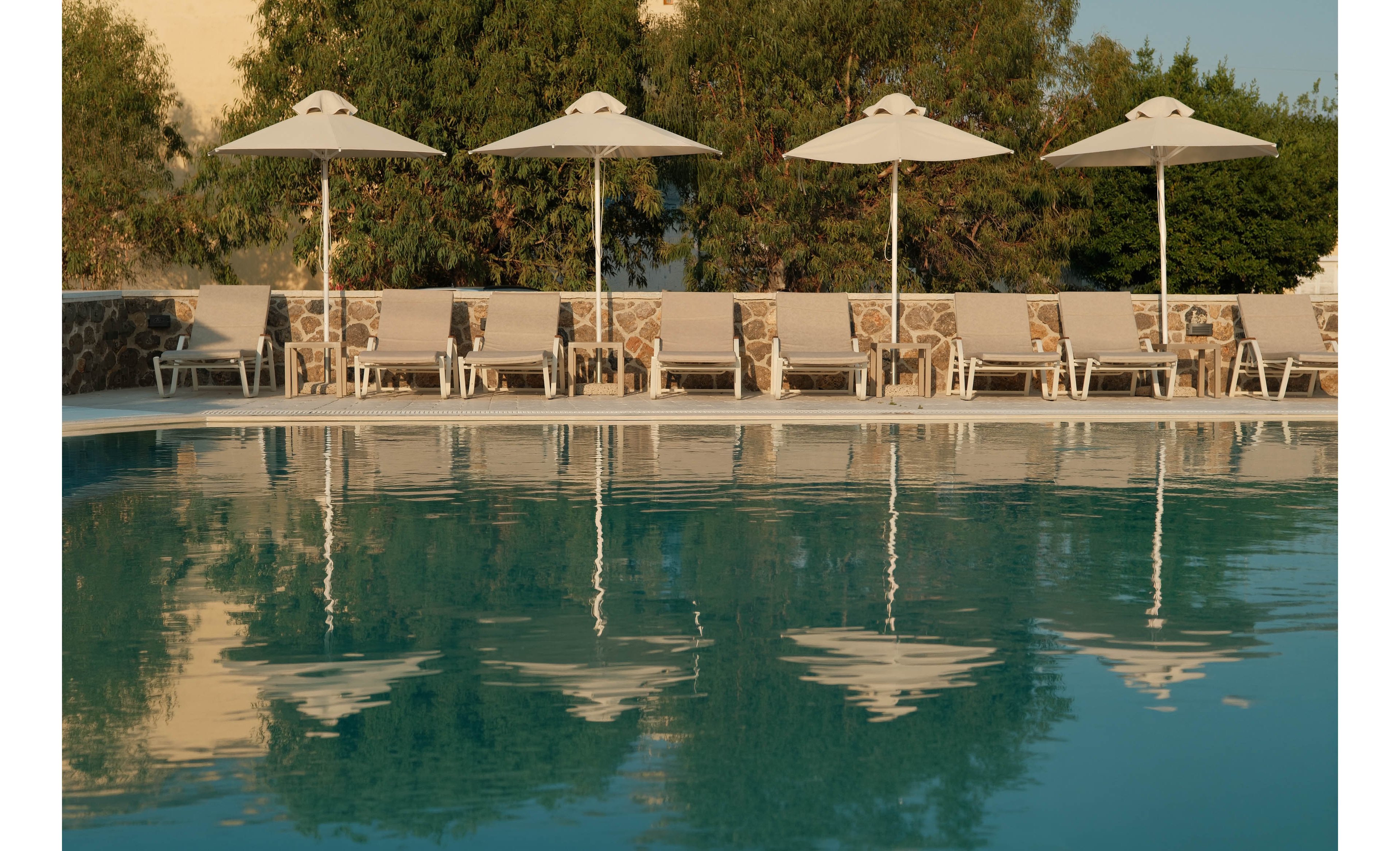 A tranquil outdoor swimming pool reflects a row of white sun loungers and umbrellas. The seating area is arranged neatly along the poolside, suggesting a resort or hotel setting. Green trees and a stone wall form a natural backdrop, adding warmth and texture to the scene. The calm water and soft lighting create a relaxed, vacation atmosphere.