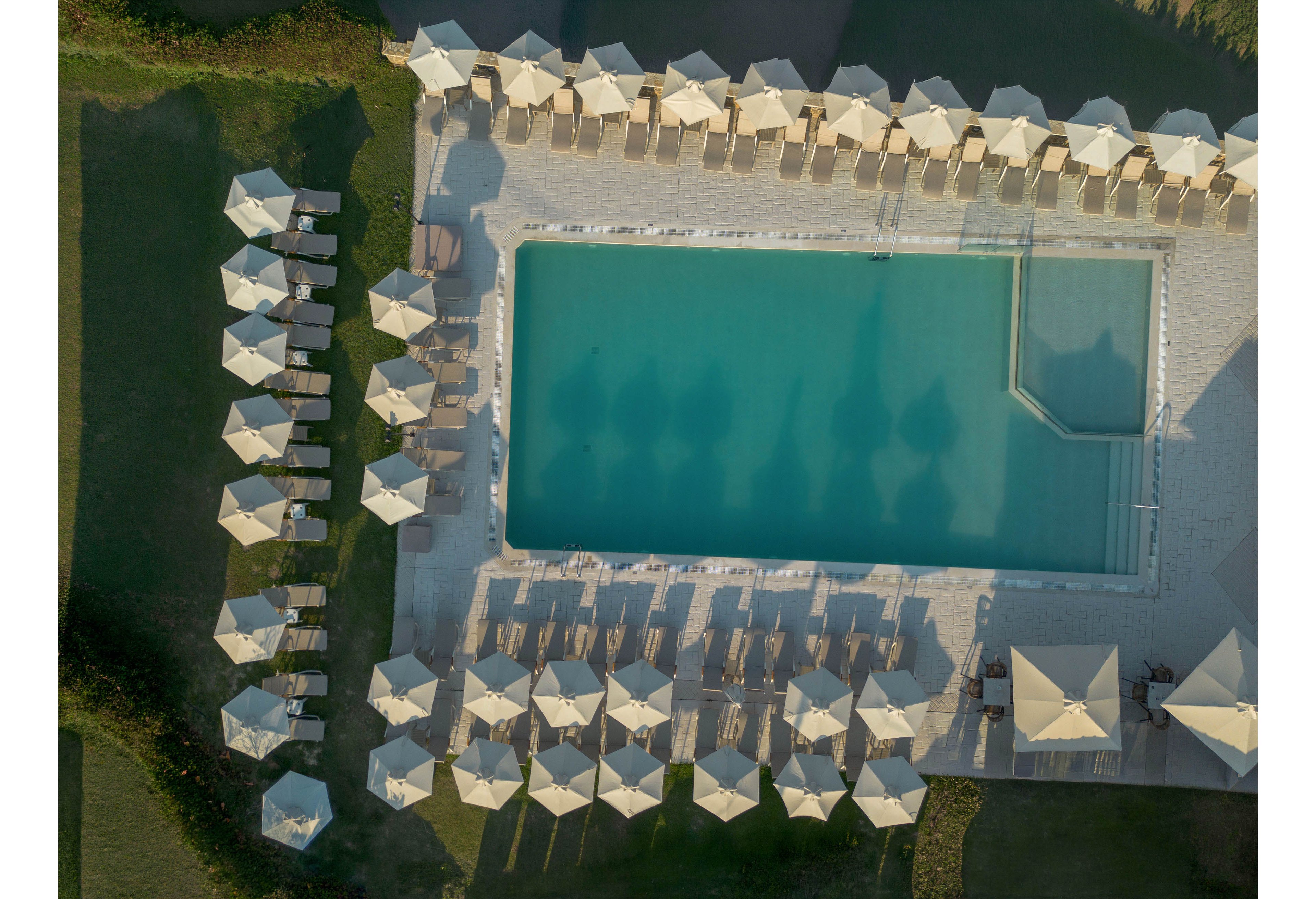 A rectangular outdoor swimming pool is photographed from above, surrounded by neatly arranged sun loungers and umbrellas. The setting appears to be a resort or hotel, with the pool bordered by a tiled deck and adjacent green lawn. Shadows of the umbrellas fall across the water, creating a symmetrical and orderly pattern. The overall scene suggests a calm, sunny day and a relaxing vacation atmosphere.